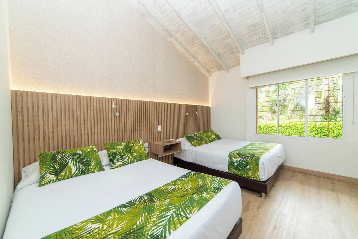 A hotel room with two beds, a wooden headboard, a window with a view of greenery, and a shelf with a book.