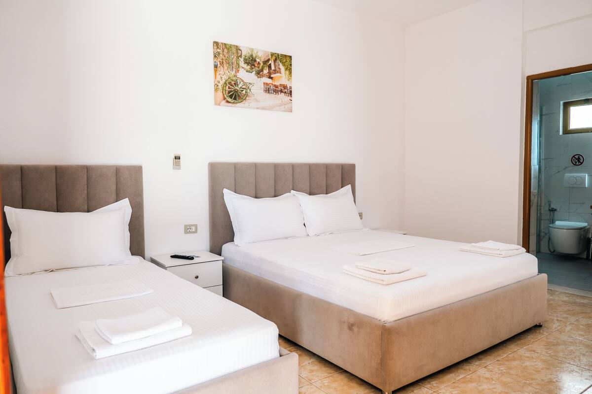 A hotel room with two beds, a nightstand, a painting on the wall, and a bathroom visible through an open door.