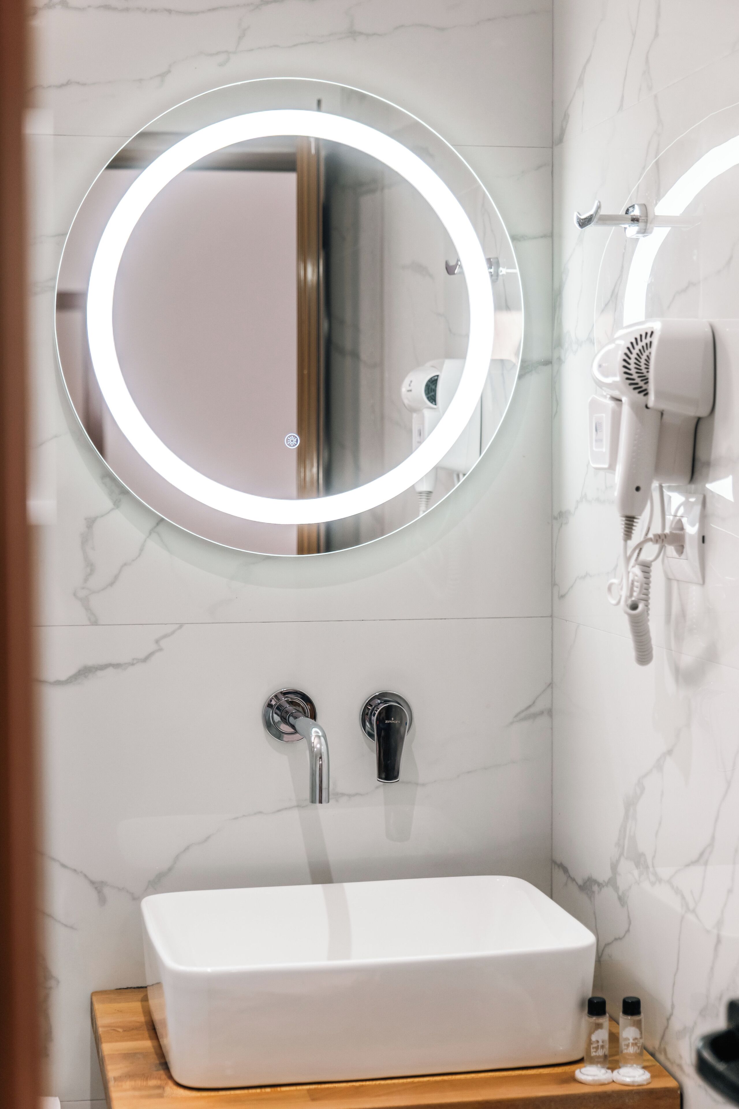 A bathroom with a white sink, a round mirror with a light, and a hairdryer on the wall.