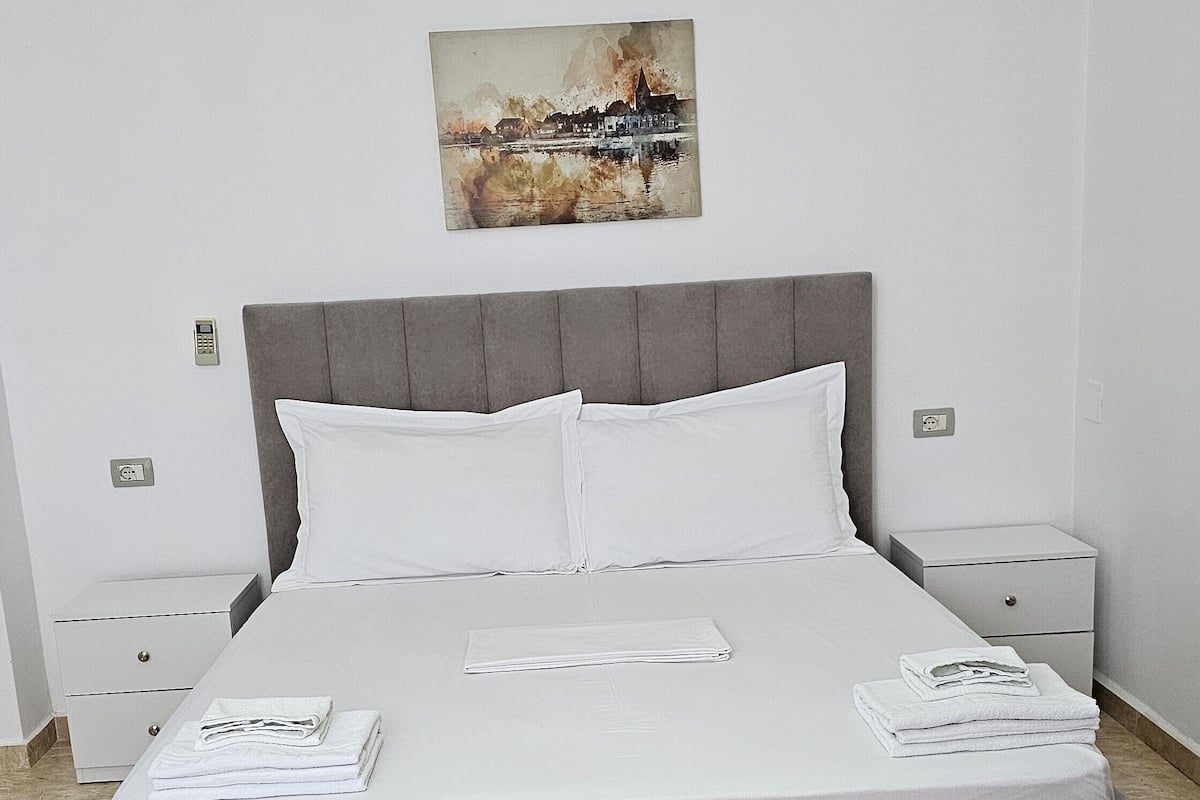 A neatly made bed with white linens, a grey headboard, and a painting above it. There are two bedside tables with lamps, a small wooden table in the foreground, and a tiled floor.