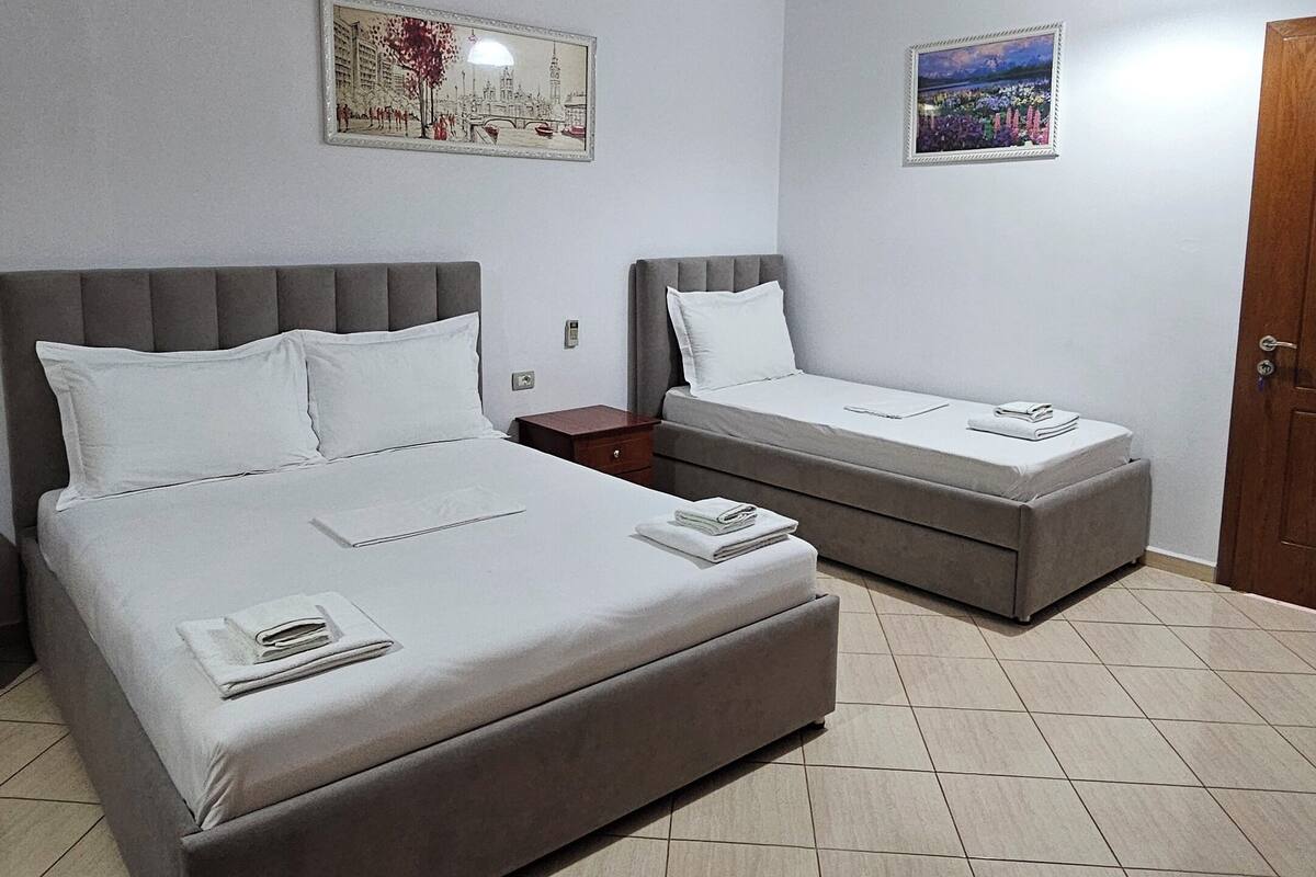 A hotel room with two beds, a nightstand, a wooden door, and two wall-mounted paintings.