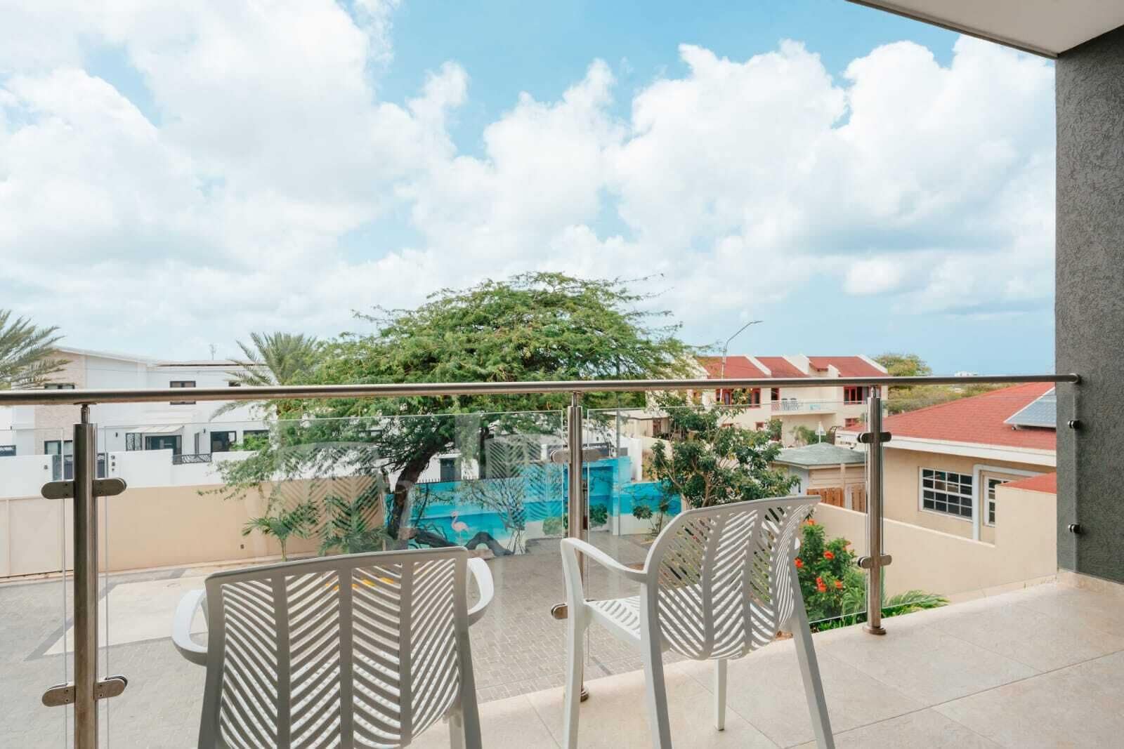 Un balcón con dos sillas, barandilla de cristal y vistas a edificios y árboles.