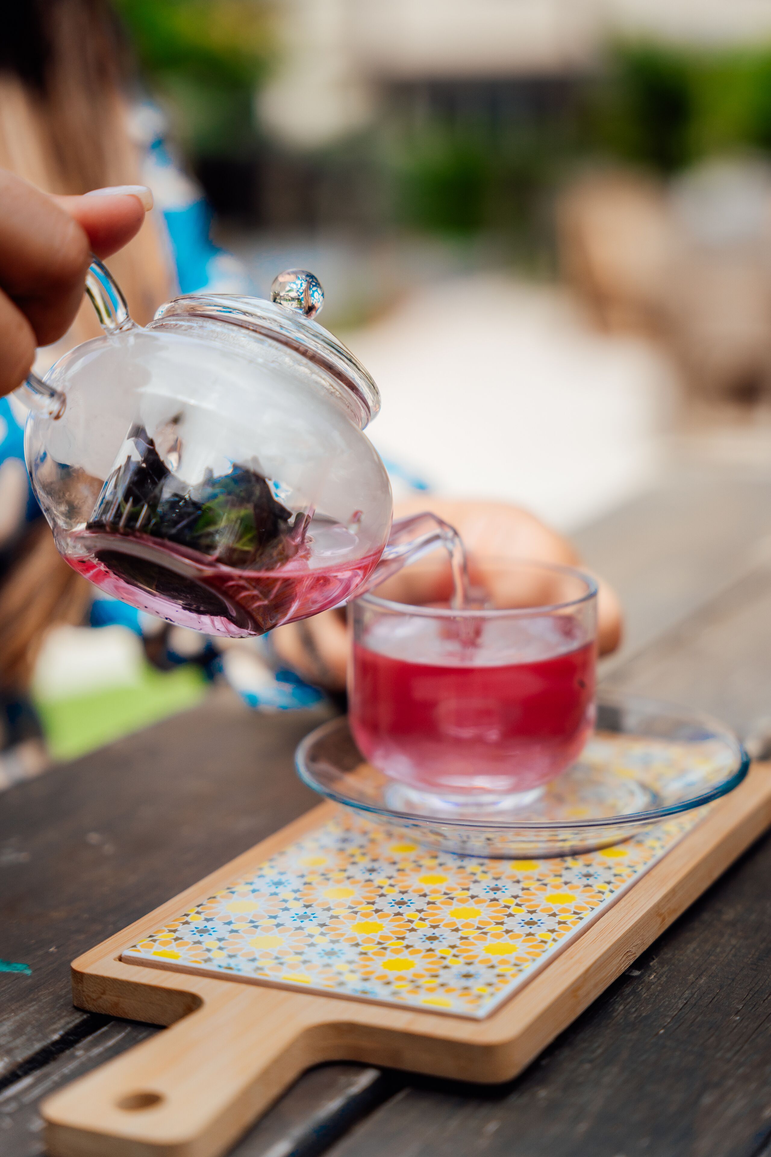 A clear glass teapot pouring tea into a glass cup on a wooden tray with a patterned coaster.