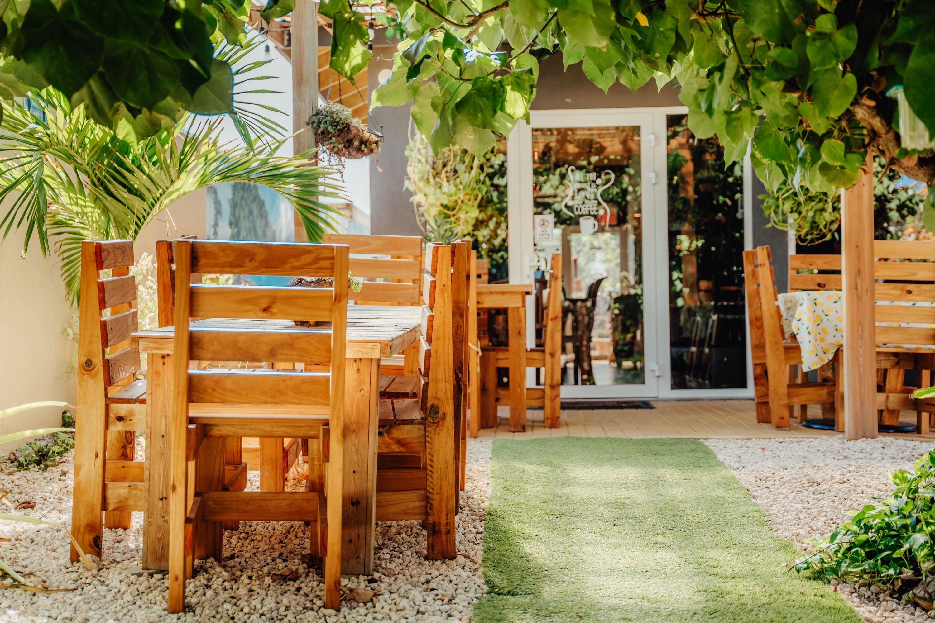 A wooden outdoor seating area with tables and chairs, surrounded by greenery and a gravel path.