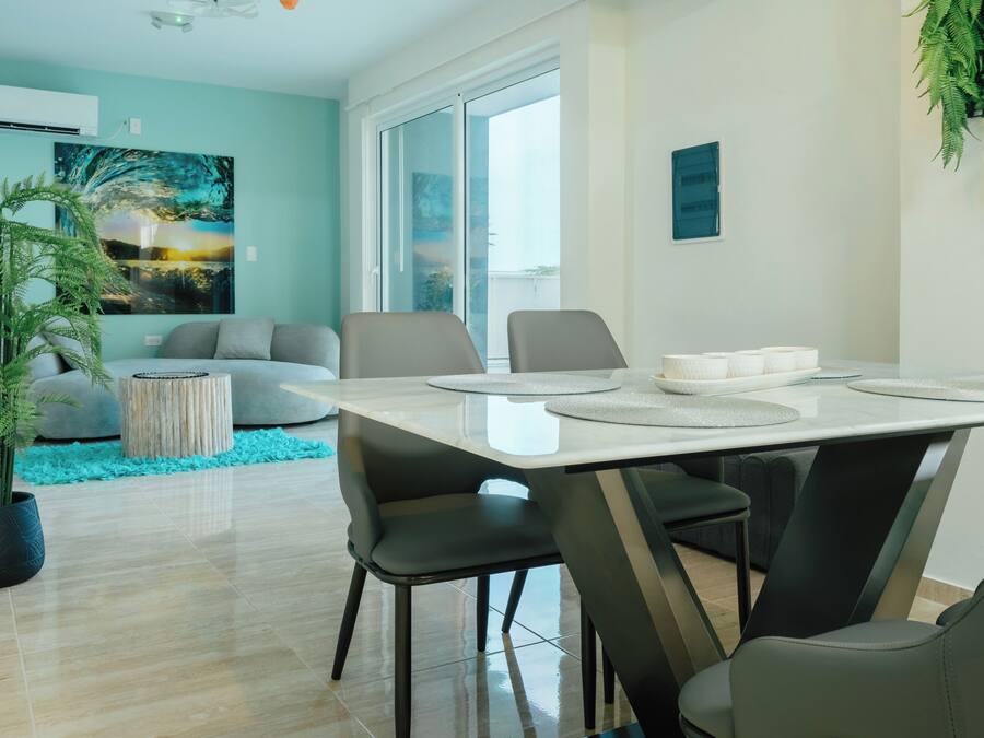 Un espace repas moderne avec une table dressée pour quatre personnes, entourée de chaises, dans un salon comprenant un canapé et une grande fenêtre.