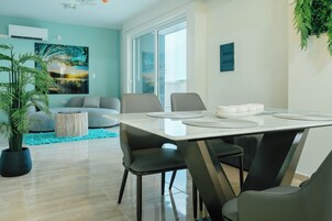 Un comedor moderno con una mesa dispuesta para cuatro personas, rodeada de sillas, en una sala de estar con un sofá y un ventanal grande.
