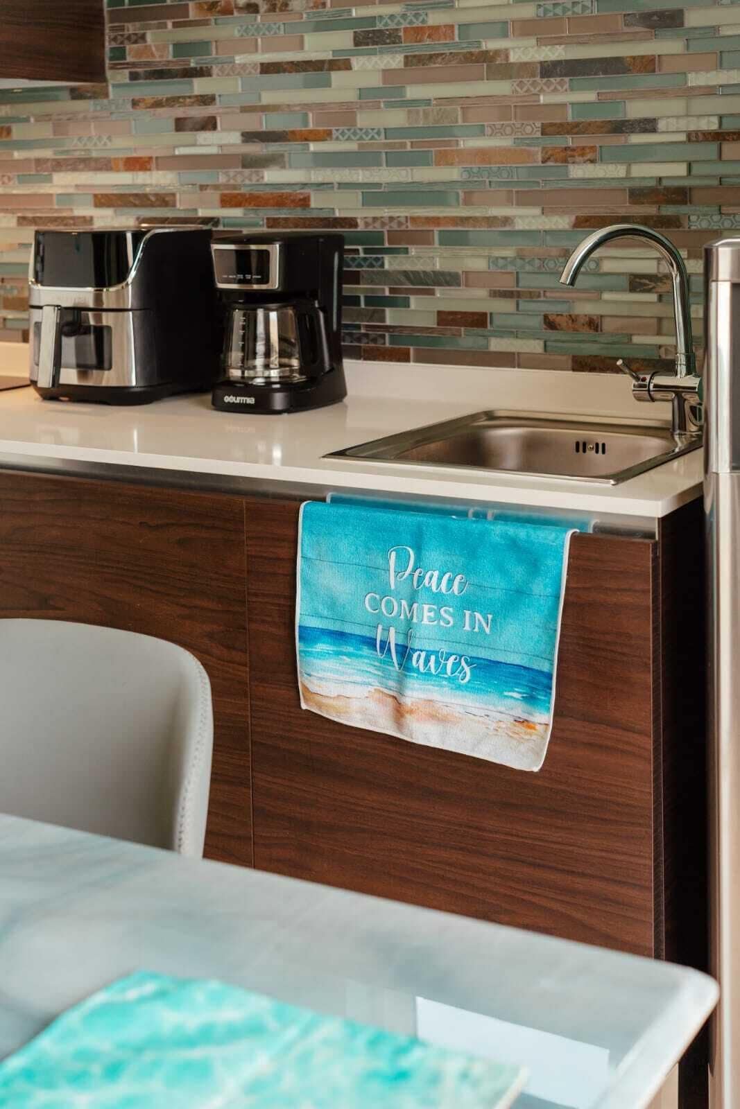 A kitchen with a tiled backsplash, a coffee maker, a sink, and a towel with a beach scene.