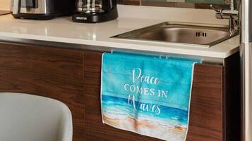 A kitchen with a tiled backsplash, a coffee maker, a sink, and a towel with a beach scene.