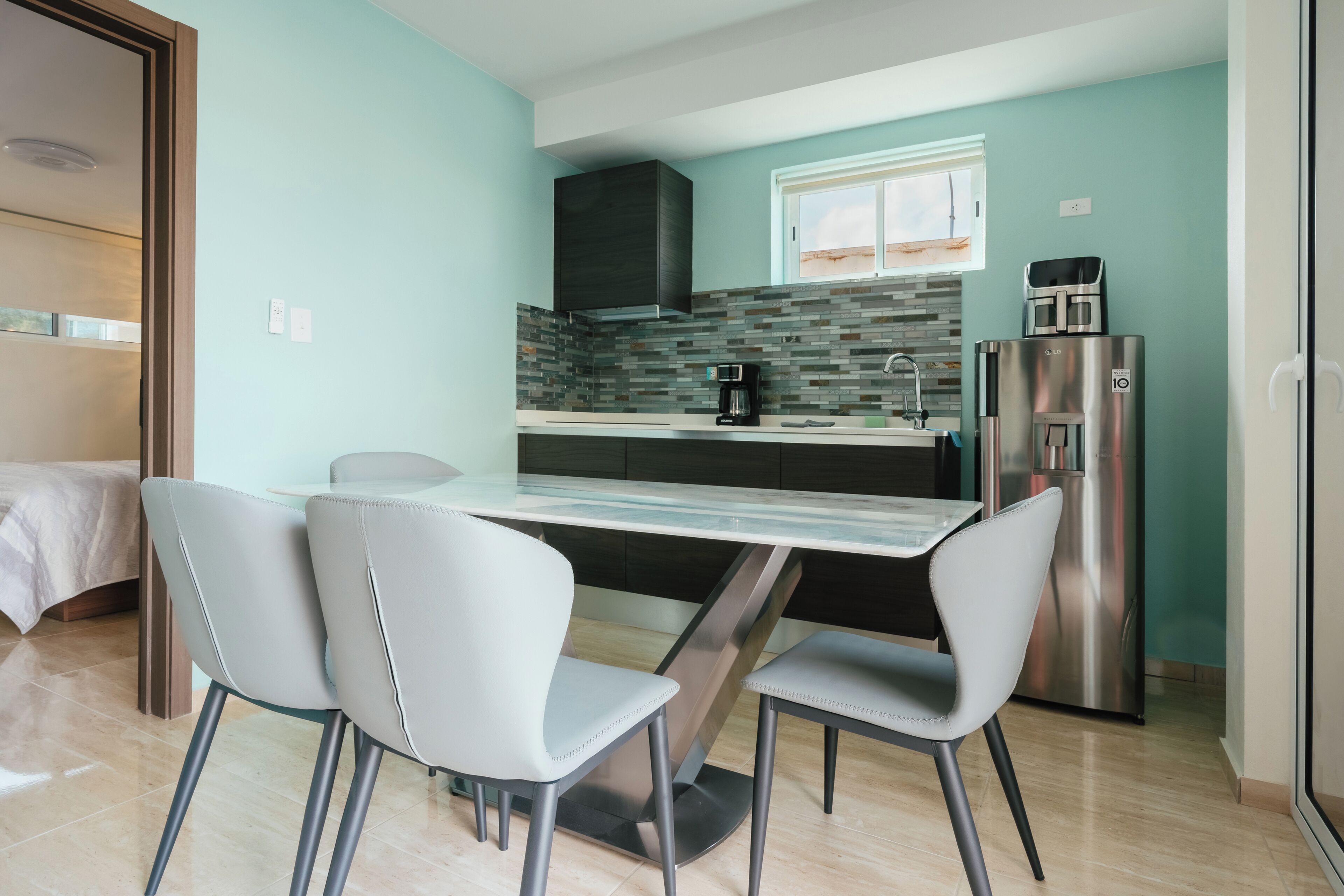A modern kitchen with a glass dining table, two chairs, a coffee maker, a refrigerator, and a window with blinds.