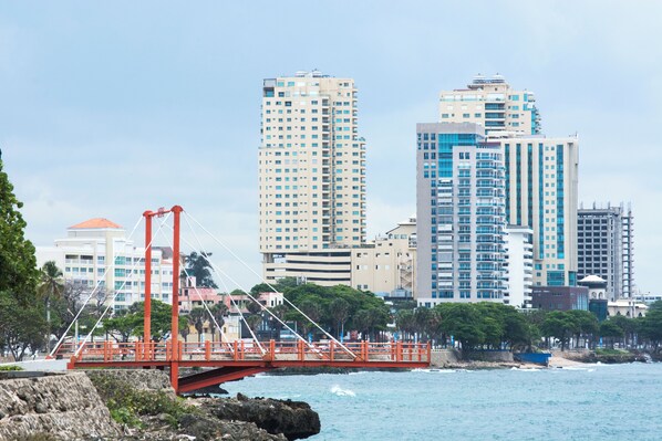 A coastal cityscape with a prominent red suspension bridge, high-rise buildings, and a rocky shoreline. - Malecon Rooms & Hotel (Santo Domingo)