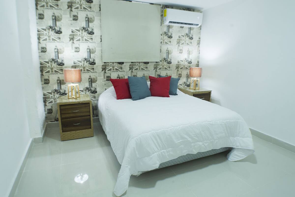 Une chambre à coucher avec un grand lit, deux tables de chevet, une commode, un climatiseur fixé au mur et un papier peint à motifs.