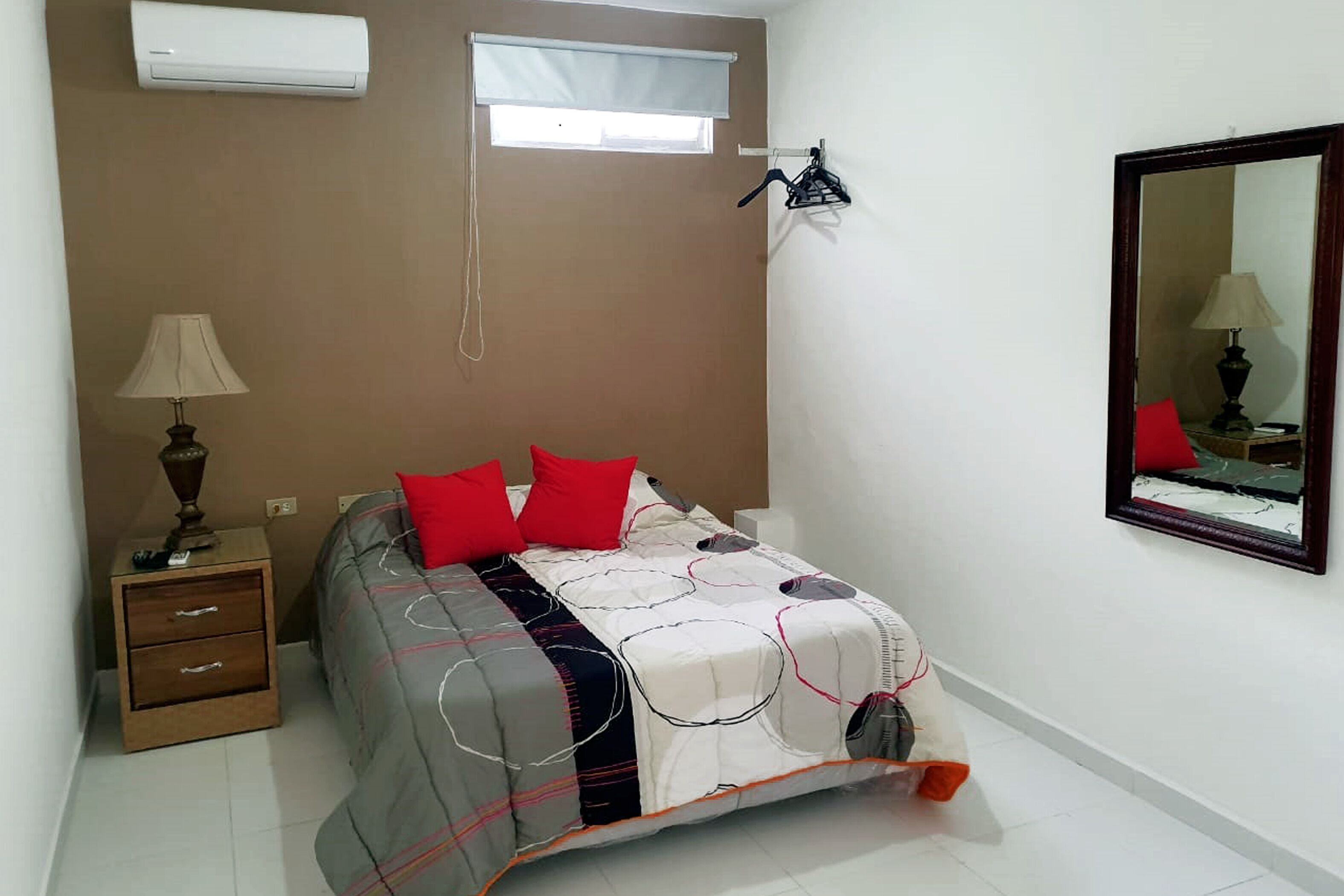Ein Schlafzimmer mit einem Bett, einem Nachttisch, einer Lampe, einem Fenster, einer Klimaanlage und einem Spiegel.