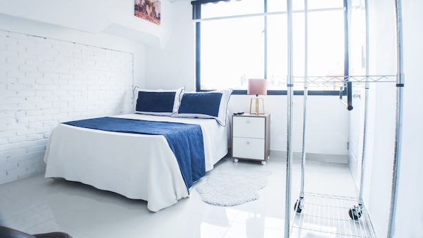 Une chambre moderne avec un mur en briques blanches, un grand lit avec une literie bleue, une table de chevet avec une lampe et une grande fenêtre.