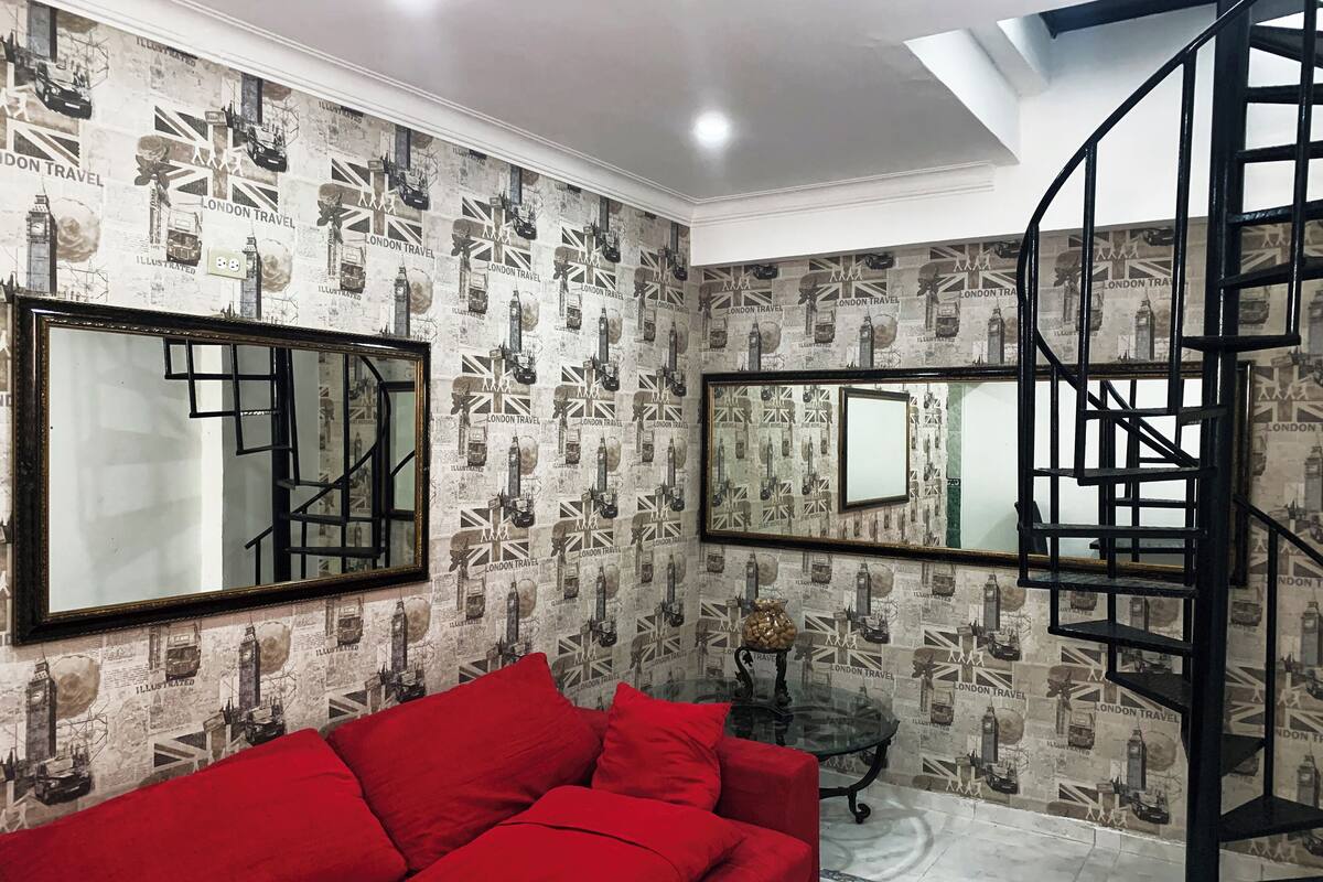 Une pièce avec un canapé rouge, deux grands miroirs, un escalier en colimaçon et un papier peint orné d’illustrations sur le thème des voyages à Londres.