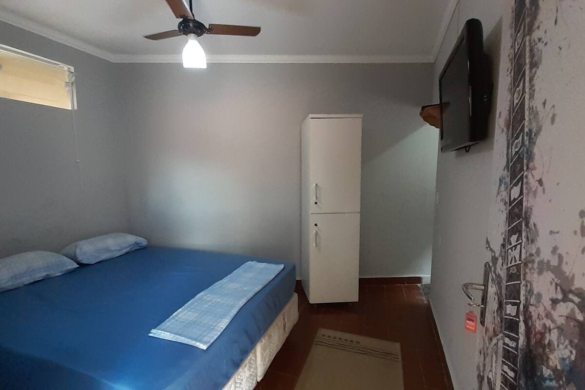 A bedroom with a bed, a ceiling fan, a wardrobe, and a television on the wall.