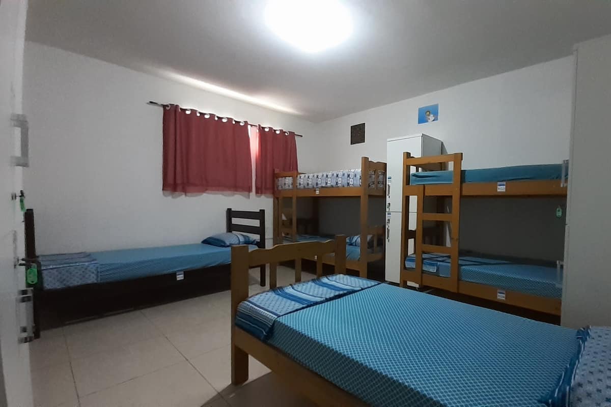 A dormitory room with bunk beds, a small refrigerator, and a window with red curtains.