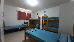 A dormitory room with bunk beds, a small refrigerator, and a window with red curtains.