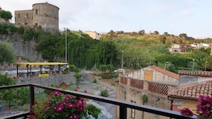 A balcony with a view of a historic stone building, a paved area with a yellow structure, and a hillside with greenery. - Giò Rooms (Scalea)