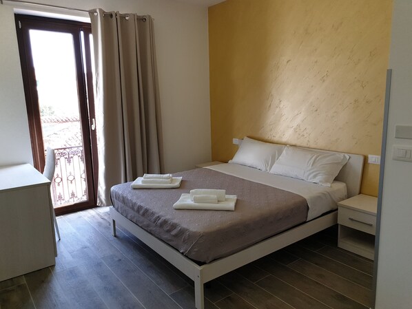 Un dormitorio ordenado con cama, mesitas de noche y vistas a través de una ventana con cortinas.