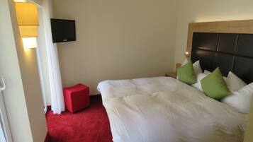 A hotel room with a bed, a red ottoman, a television, and a wall-mounted lamp.