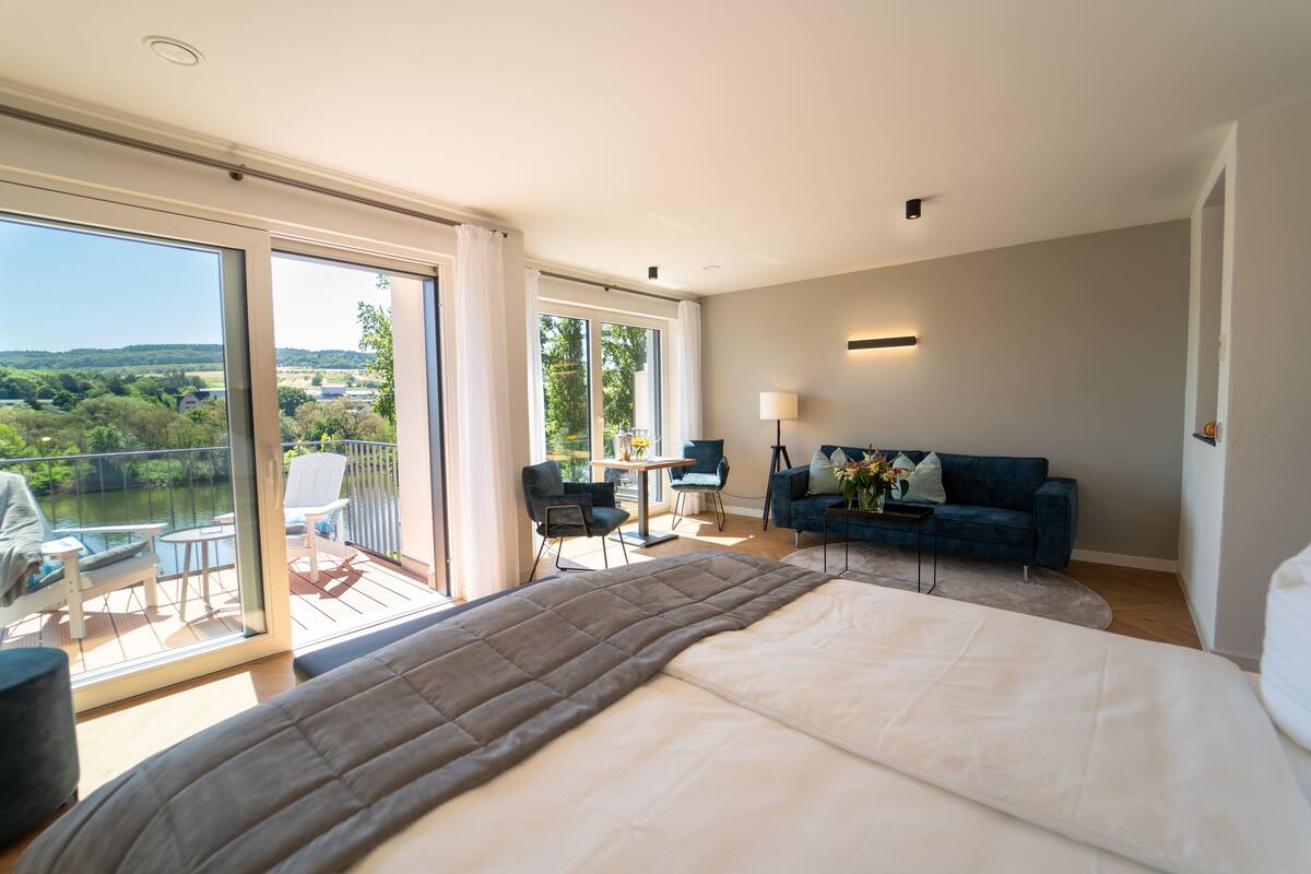 A spacious bedroom with a large bed, a balcony with a view of greenery, a seating area with a table and chairs, and a blue sofa.
