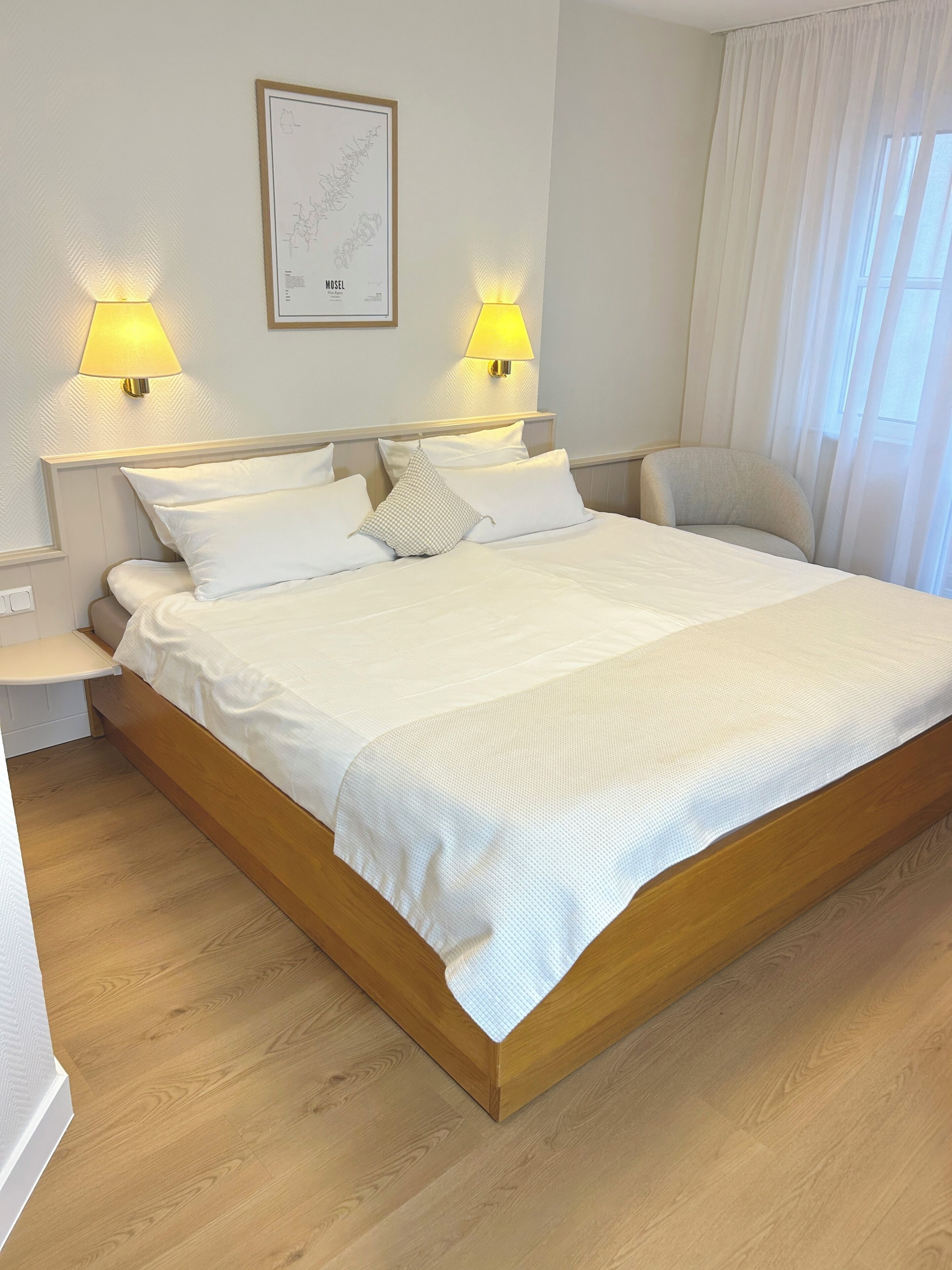 Ein ordentlich bezogenes Bett mit weißen Leinentüchern, zwei Wandlampen und eine gerahmte Karte an der Wand.