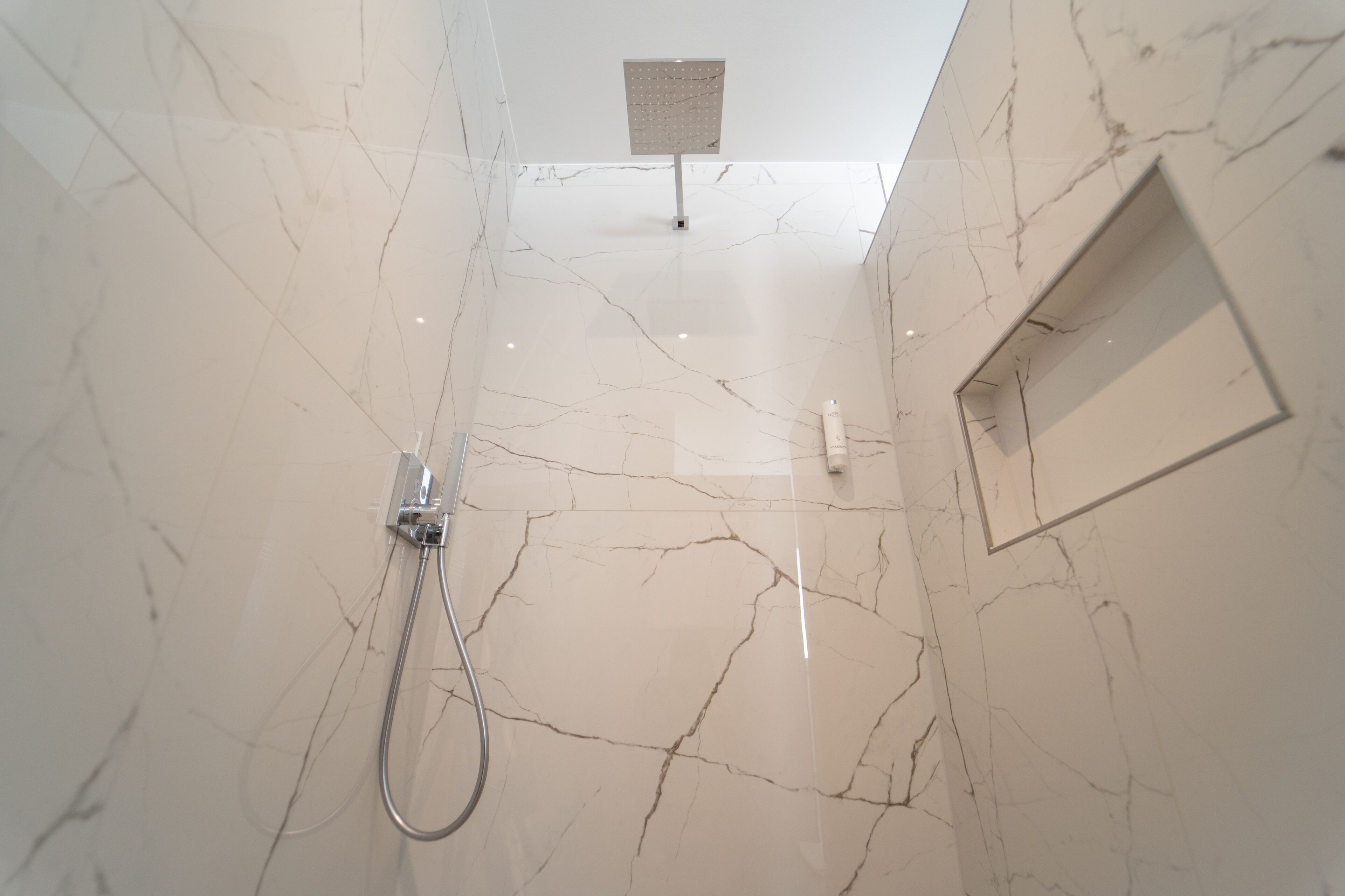 Ein modernes Badezimmer mit Marmorwänden, einer Dusche mit Handbrause und einer fest installierten, rechteckigen Duschköpfe.