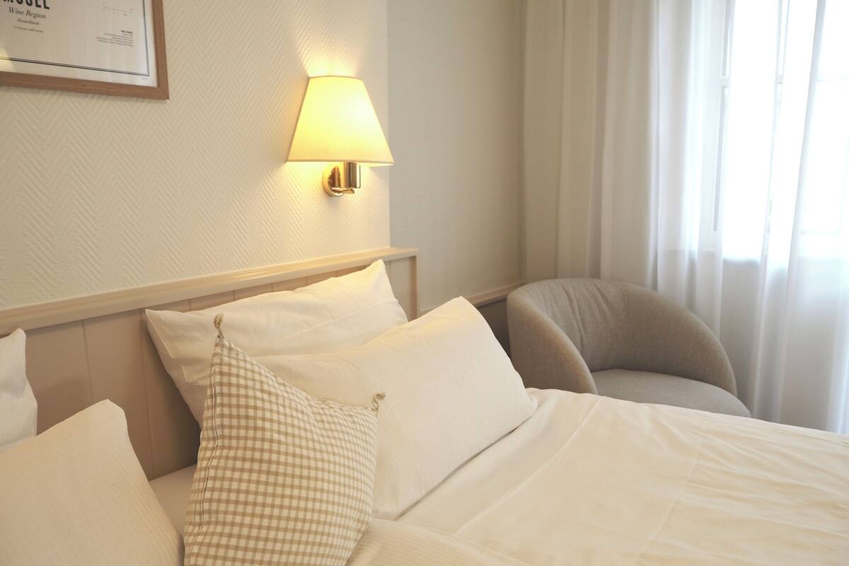 A hotel room with a bed, pillows, a lamp, a chair, and a window with curtains.