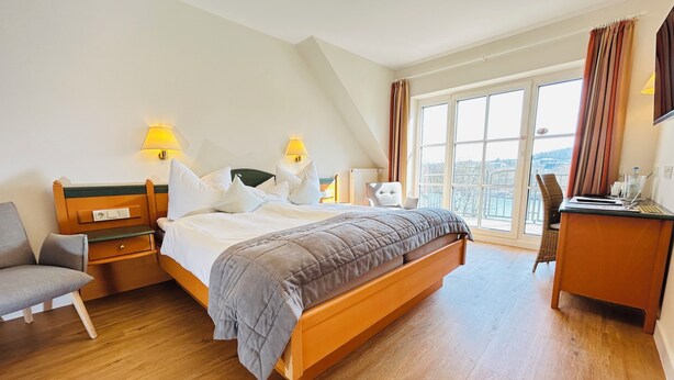 Quarto de hotel moderno com uma cama grande, uma escrivaninha e uma cadeira. Há duas luminárias de cabeceira e vista para a cidade pela janela.