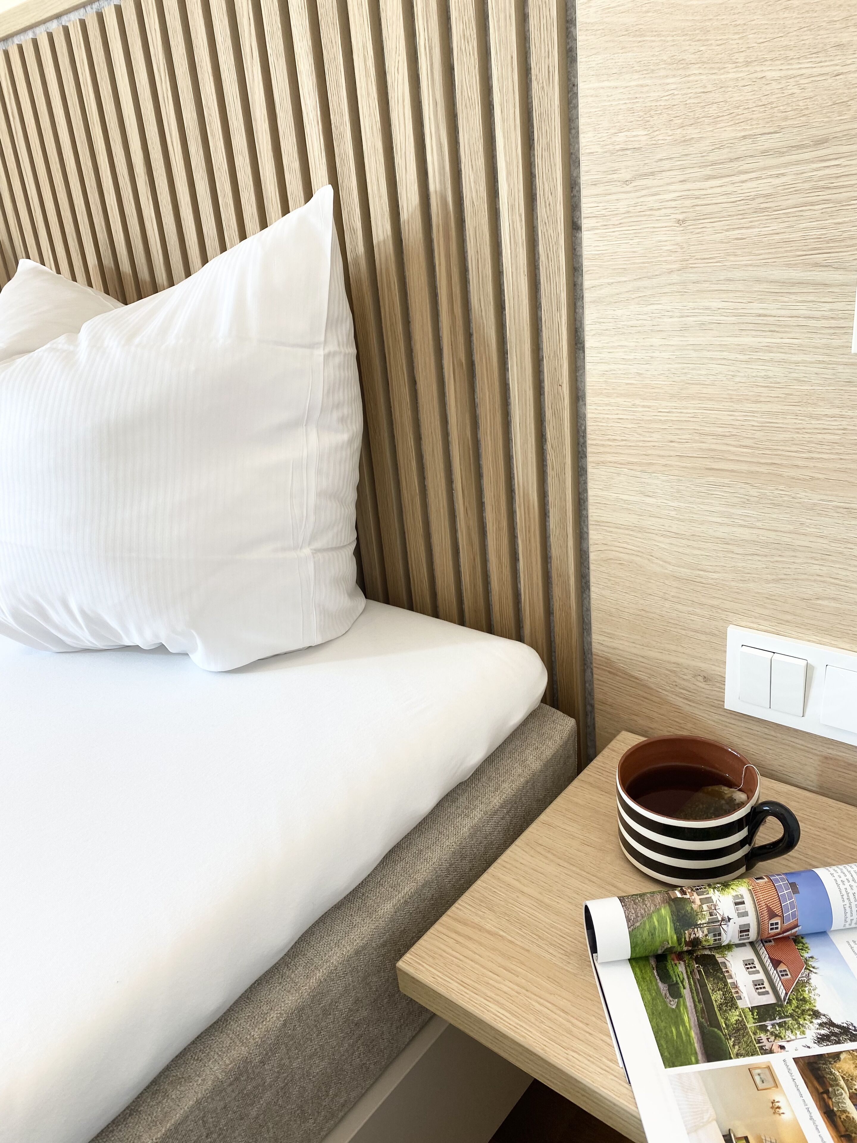 A neatly made bed with white linens and a pillow, a wooden bedside table with a mug and a magazine, and a light switch on the wall.