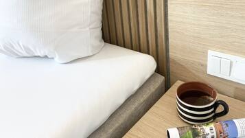 A neatly made bed with white linens and a pillow, a wooden bedside table with a mug and a magazine, and a light switch on the wall.