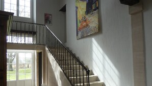 Un escalier avec une rampe noire menant à un deuxième étage, un grand tableau accroché au mur et une vue sur l’extérieur par une fenêtre.