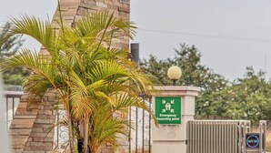 A building entrance with a stone archway, a palm tree planter, and a green emergency assembly point sign. - The Crest Lodge (Lusaka)