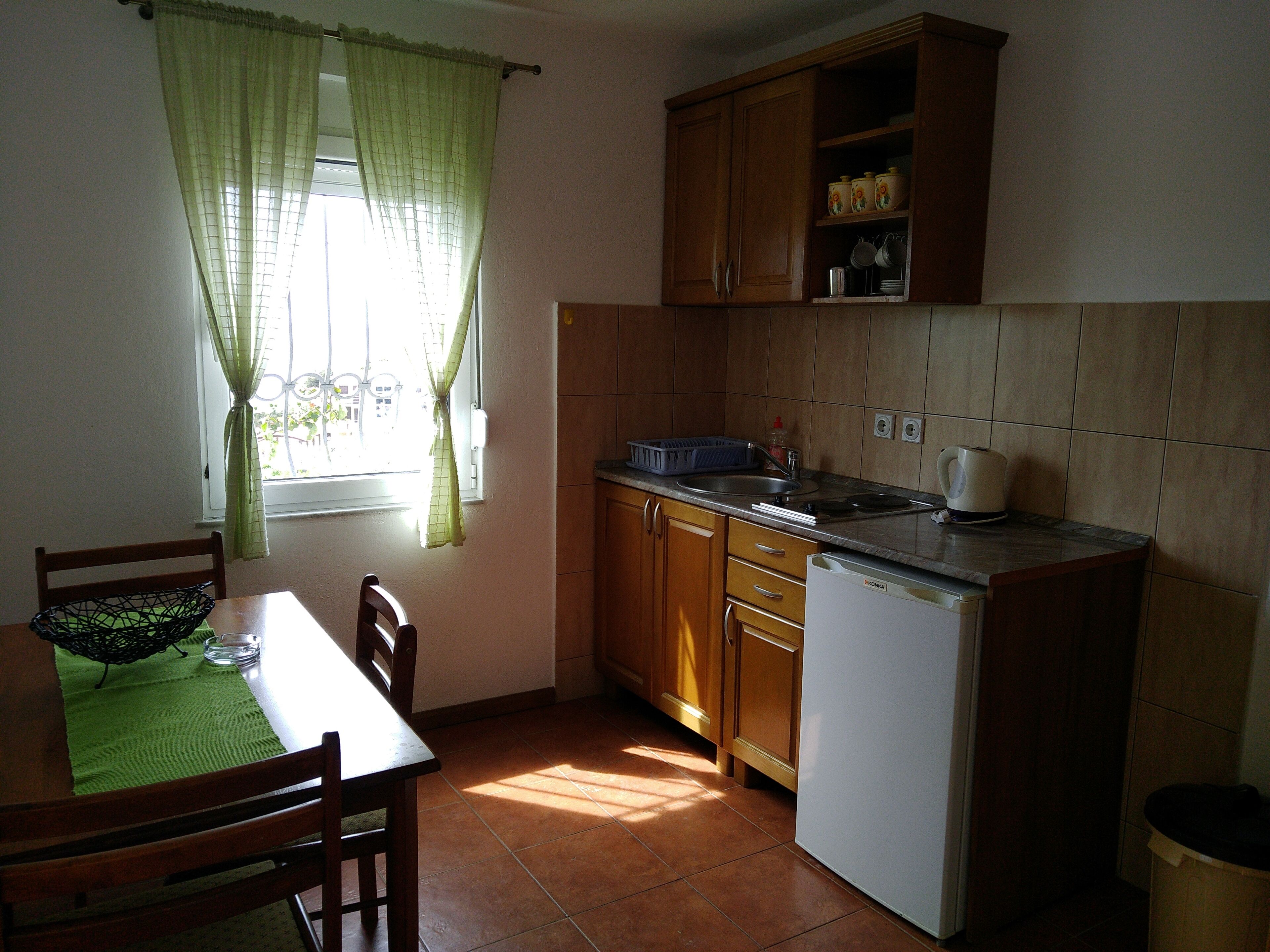 A compact kitchen with wooden cabinets, a sink, and a refrigerator. There is a window with curtains, a dining table with chairs, and a green mat.