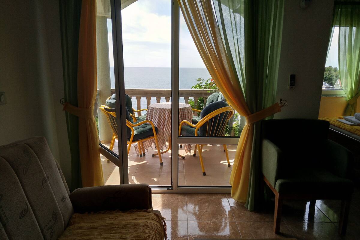 A room with a sofa, a chair, a small table, and a balcony with a table and chairs.