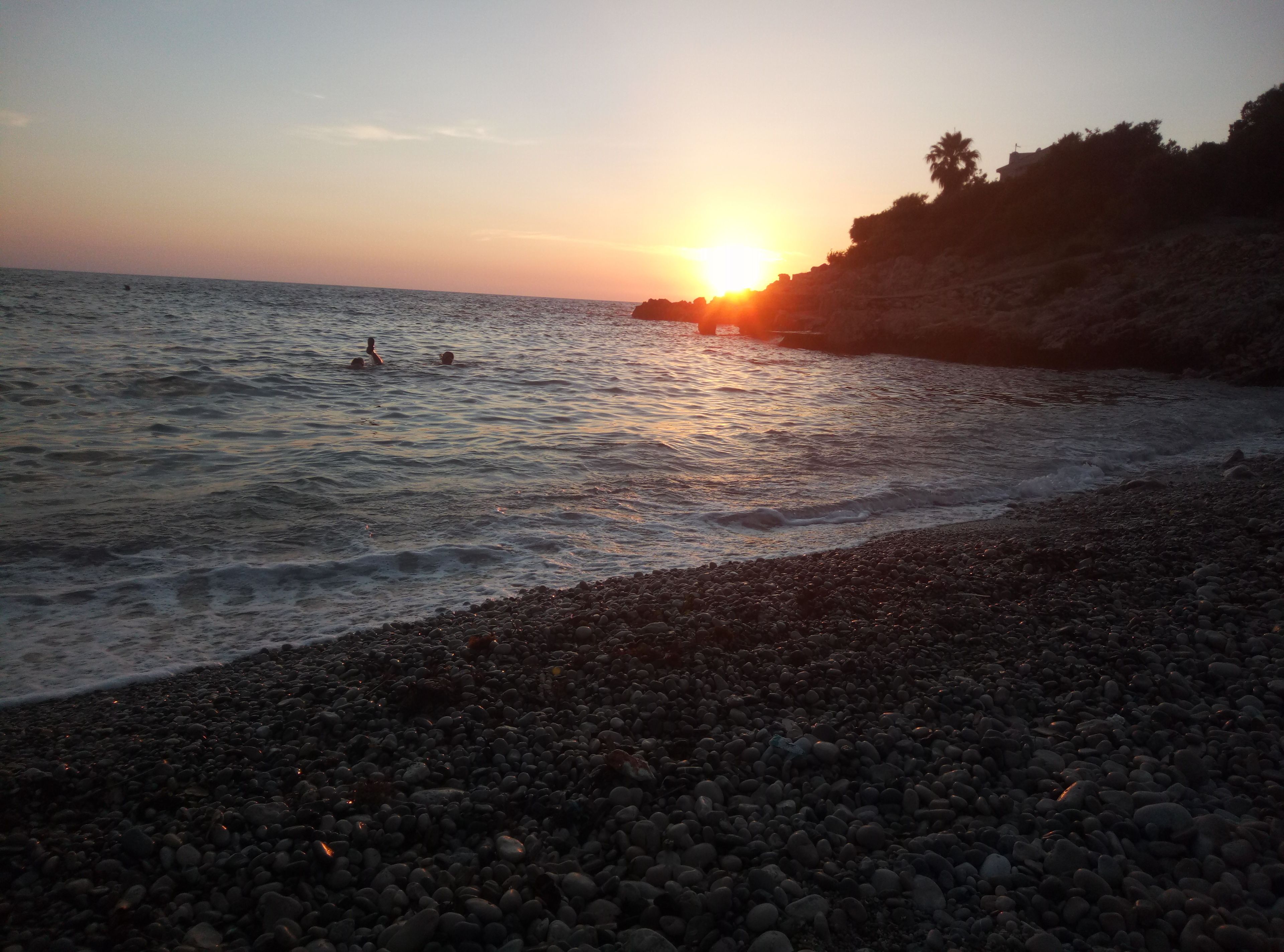 A beach with pebbles and calm water, a sunset in the background, and a few people swimming.