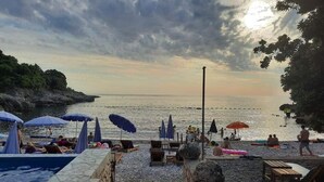 A beachside pool area with lounge chairs and umbrellas, a rocky shoreline, and a view of the sea. - Majero Apartments (Utjeha)