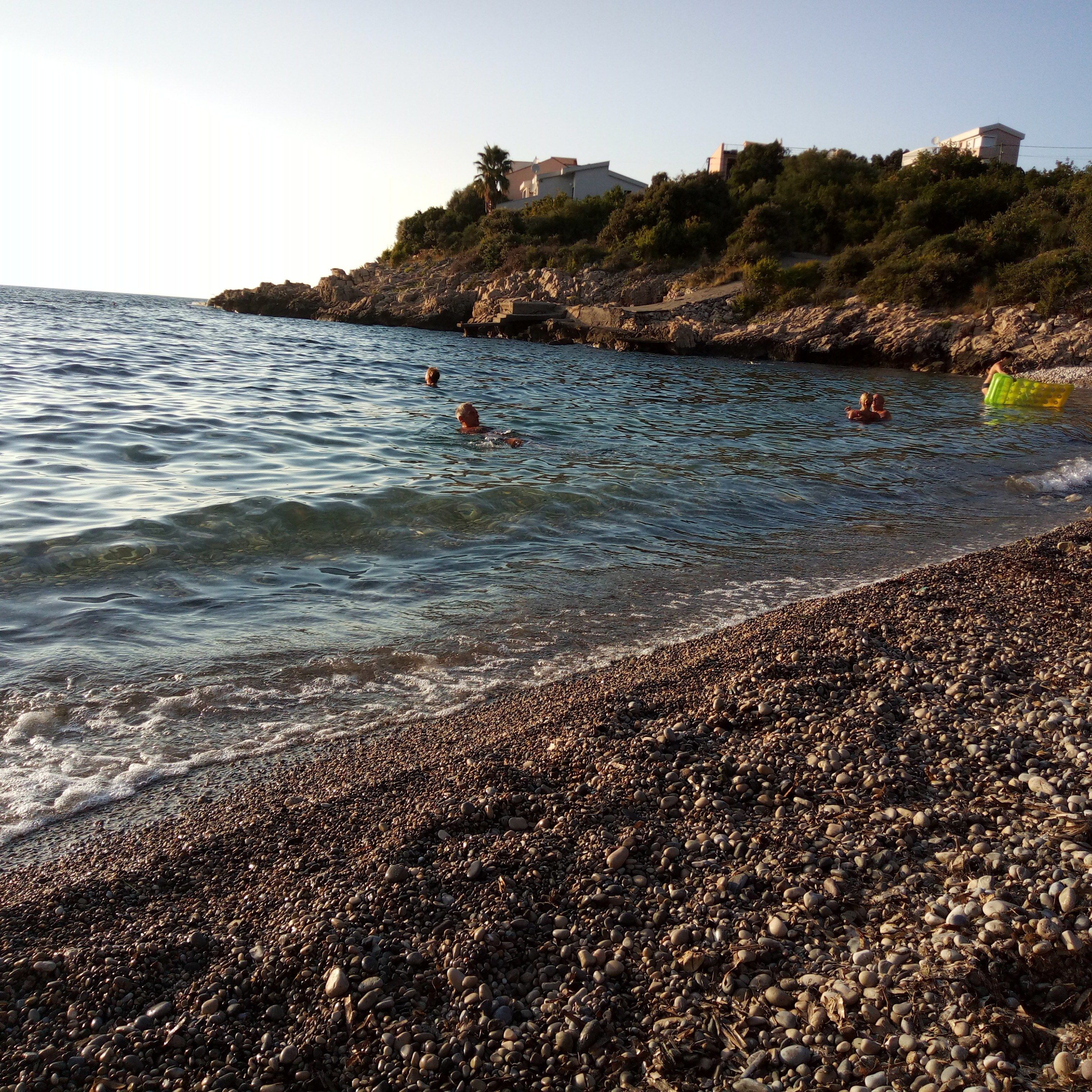 A pebbled beach with clear water, a rocky shoreline, and buildings on a hill in the background.