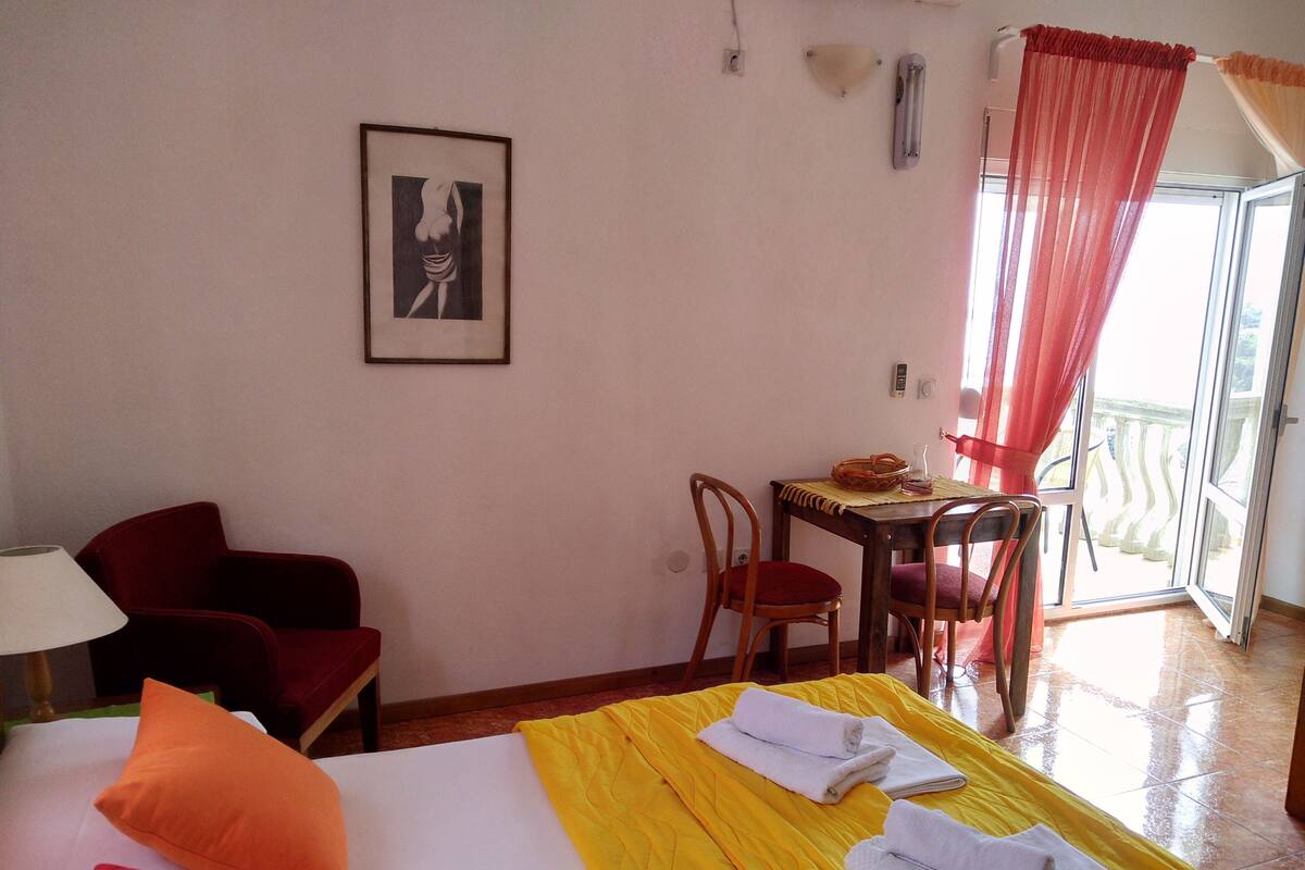 A hotel room with a bed, a desk with a chair, a red chair, a lamp, and a balcony with a view.