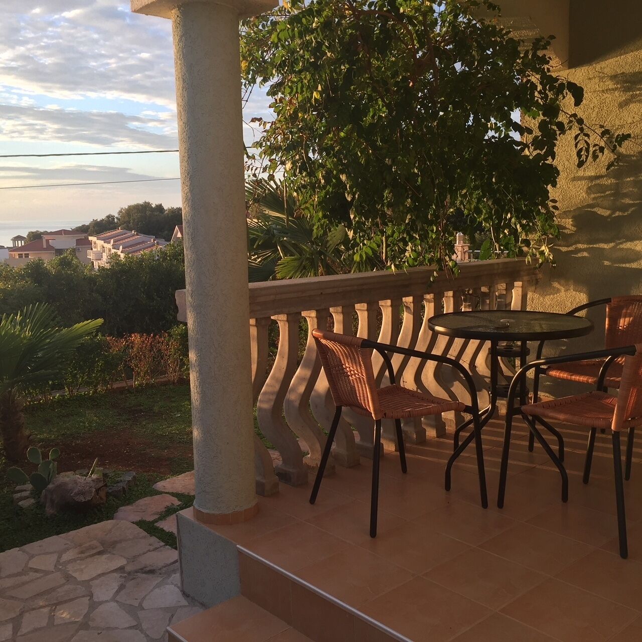 Une terrasse dotée de chaises et d’une table en osier, offrant une vue sur un jardin et, au loin, sur des maisons.