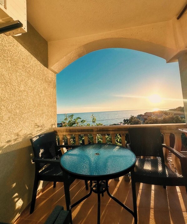 A balcony with a round table and chairs, overlooking a coastal view with the sun setting. - Majero Apartments (Utjeha)