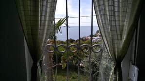 A window with metal bars and sheer curtains, overlooking a coastal landscape with greenery and buildings. - Majero Apartments (Utjeha)