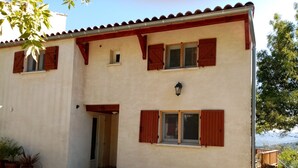 Ein zweistöckiges Gebäude mit beiger Fassade, roten Fensterläden und einem kleinen Fenster mit Leuchte darüber.