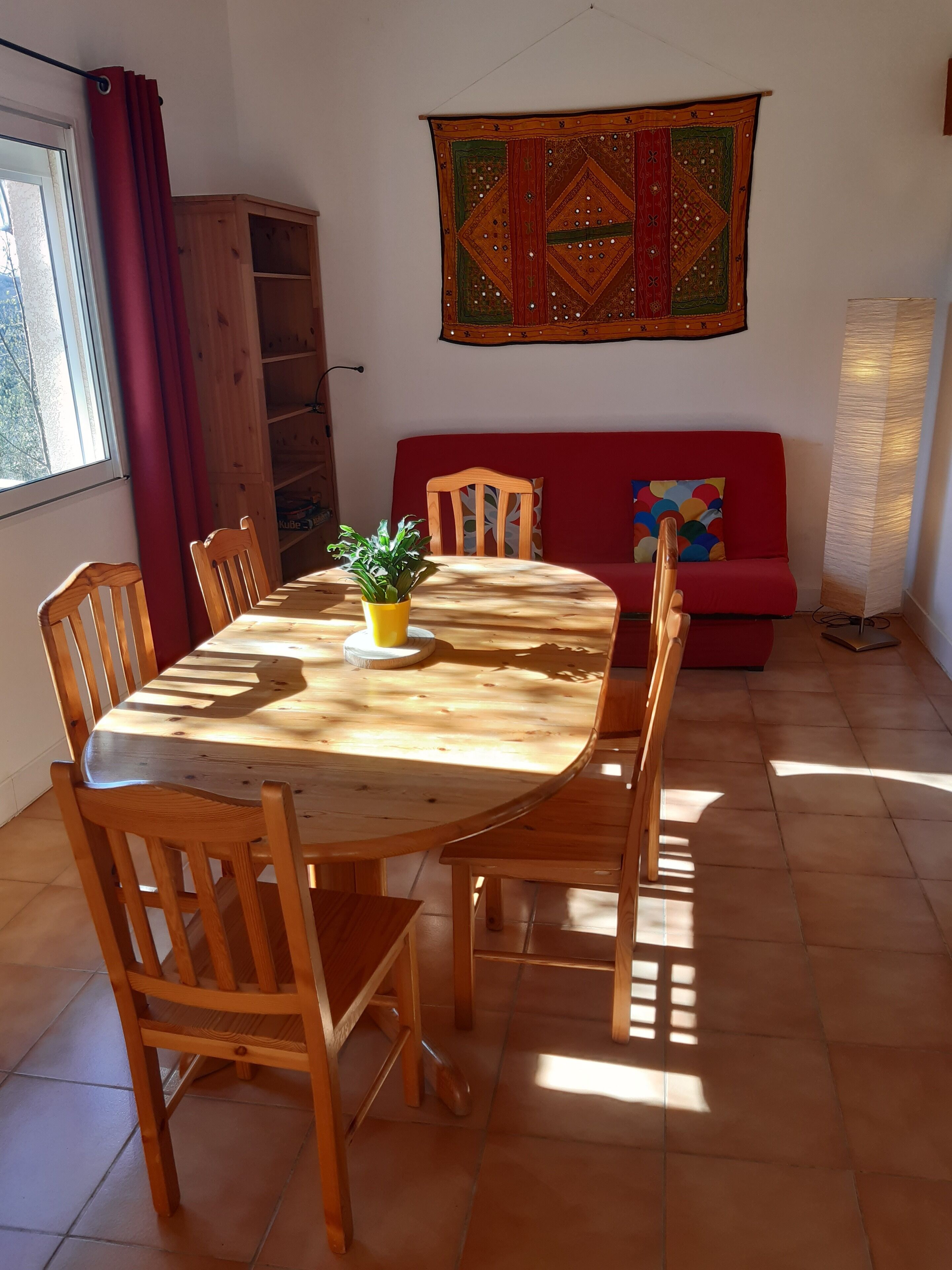 A dining area with a wooden table and chairs, a potted plant, a red sofa, and a colorful tapestry on the wall.