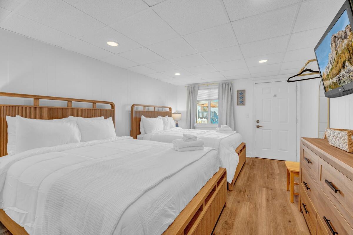 Une chambre d’hôtel avec deux lits, des têtes de lit en bois, du linge de lit blanc, une commode en bois, une télévision et une porte.