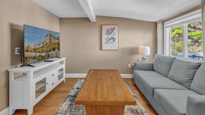 Un salon avec une table basse en bois, un canapé gris, un meuble de télévision blanc avec un téléviseur, un tableau encadré au mur et une fenêtre donnant sur un paysage.