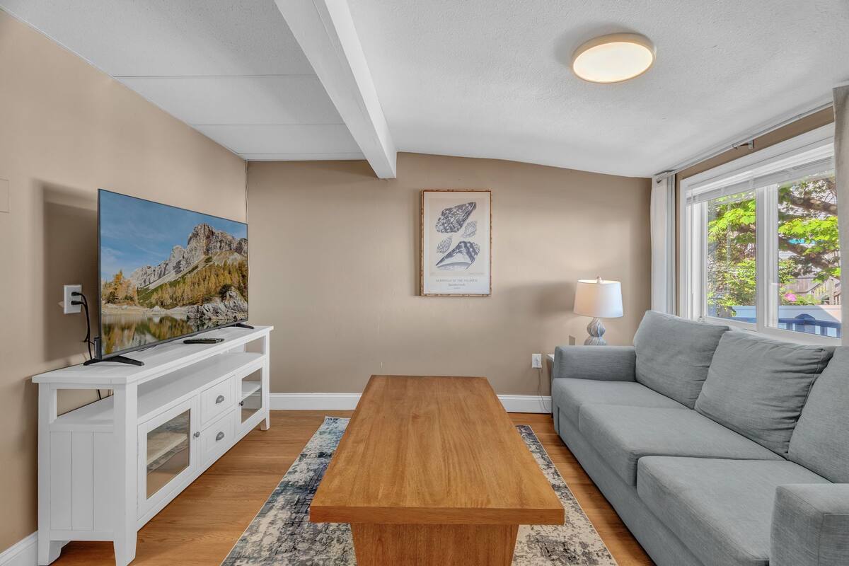 Un salon avec une table basse en bois, un canapé gris, un meuble de télévision blanc avec un téléviseur, un tableau encadré au mur et une fenêtre donnant sur un paysage.