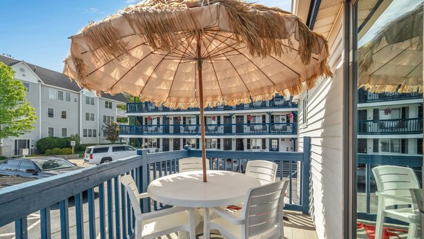 Un balcon avec une table ronde et des chaises, un parasol en paille, et une vue sur un immeuble doté de balcons.