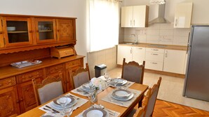 Un comedor con una mesa de madera dispuesta para cuatro personas, rodeada de sillas de madera. Hay una zona de cocina con armarios blancos y un refrigerador de acero inoxidable.