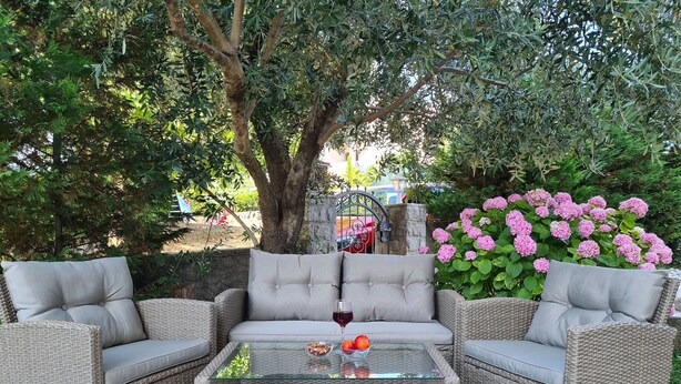 Un área de estar al aire libre con muebles de mimbre, una mesa de centro de vidrio, sillas acolchadas y un fondo de árboles y flores rosadas.